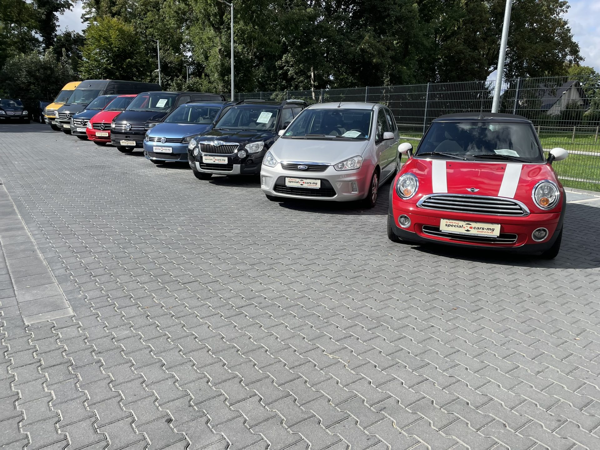 Eine Reihe verschiedener geparkter Autos, darunter Minivans und ein roter Mini Cooper, stehen auf einem gepflasterten Parkplatz mit Bäumen im Hintergrund.