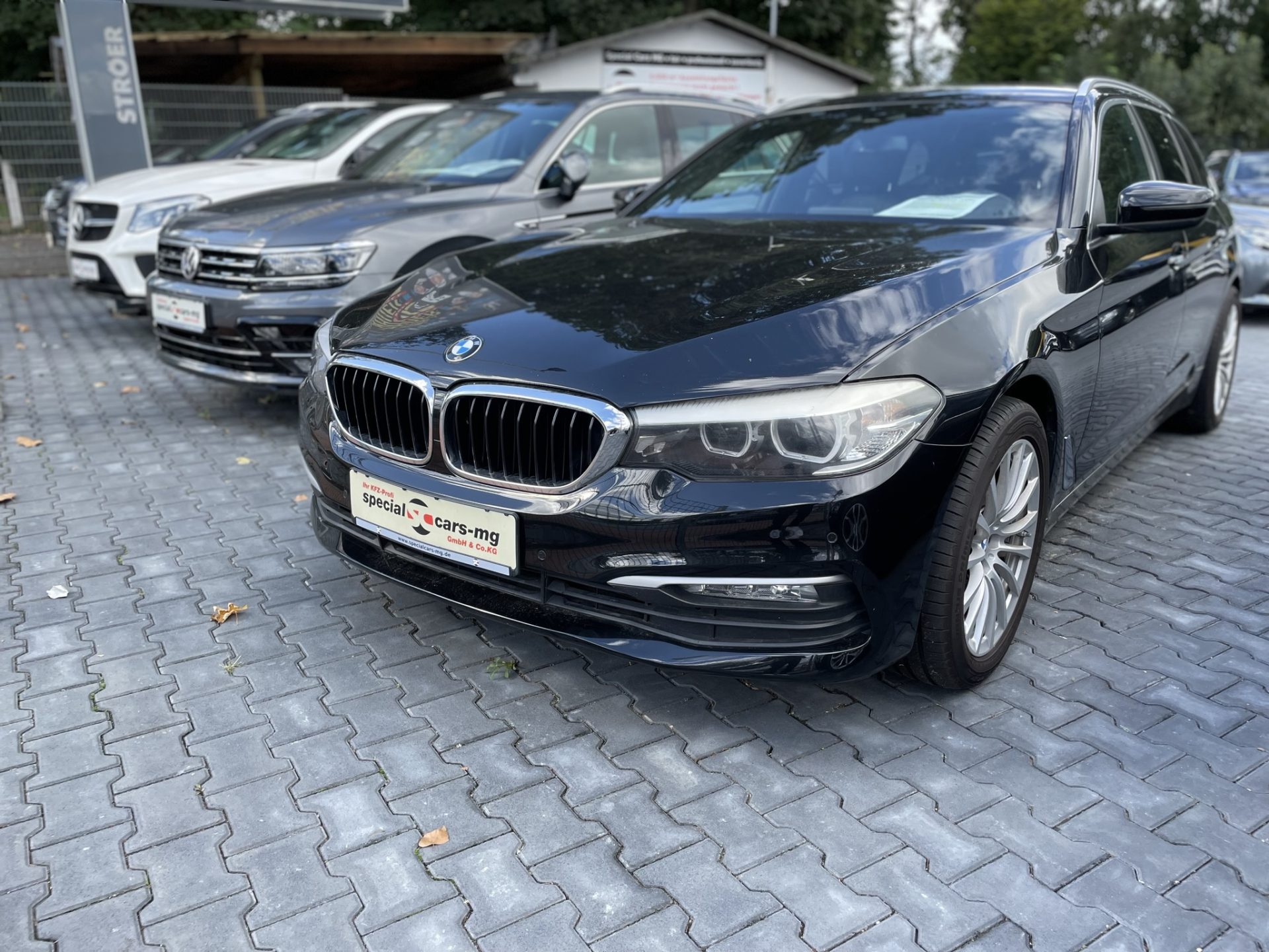 Ein schwarzer BMW steht prominent bei einem Händler, im Hintergrund stehen zwei weitere Fahrzeuge auf einem gepflasterten Parkplatz.