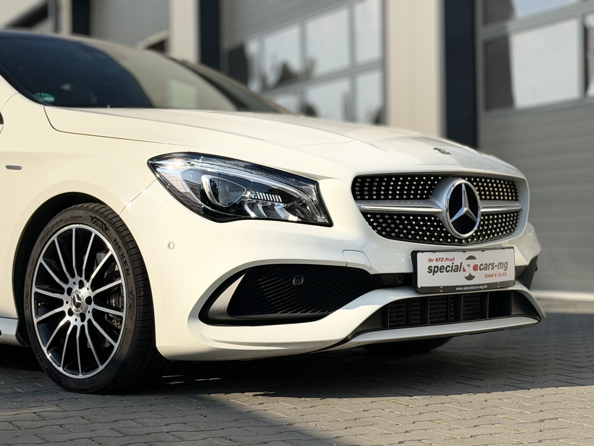 Nahaufnahme eines eleganten weißen Mercedes-Benz mit markantem Kühlergrill und sportlichen Leichtmetallfelgen, der vor einem modernen Autohaus geparkt ist.