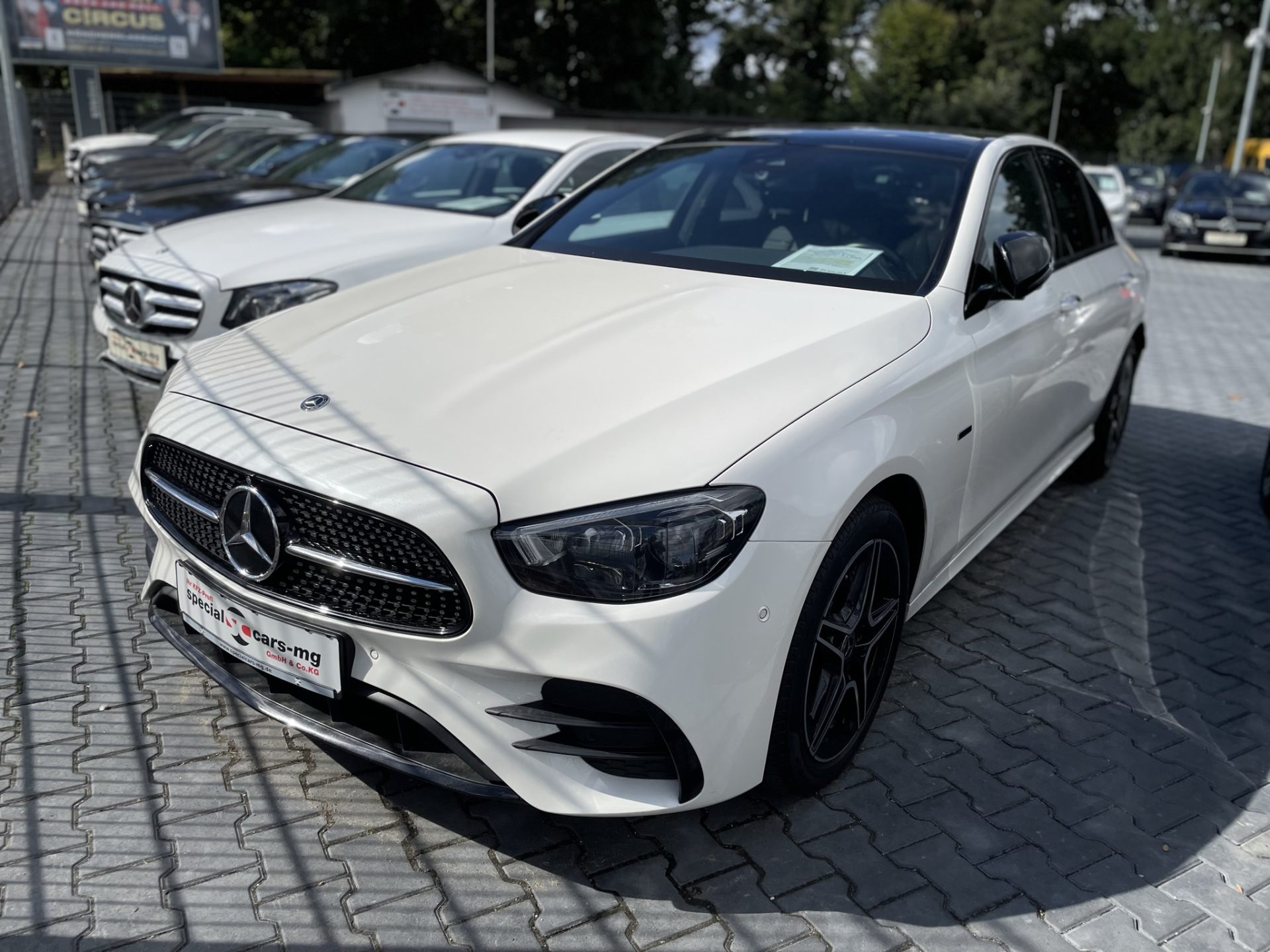 Eine elegante weiße Mercedes-Benz-Limousine, die zwischen mehreren Fahrzeugen auf einem Händlergelände geparkt ist, präsentiert ihr modernes Design und ihre sportlichen Akzente.