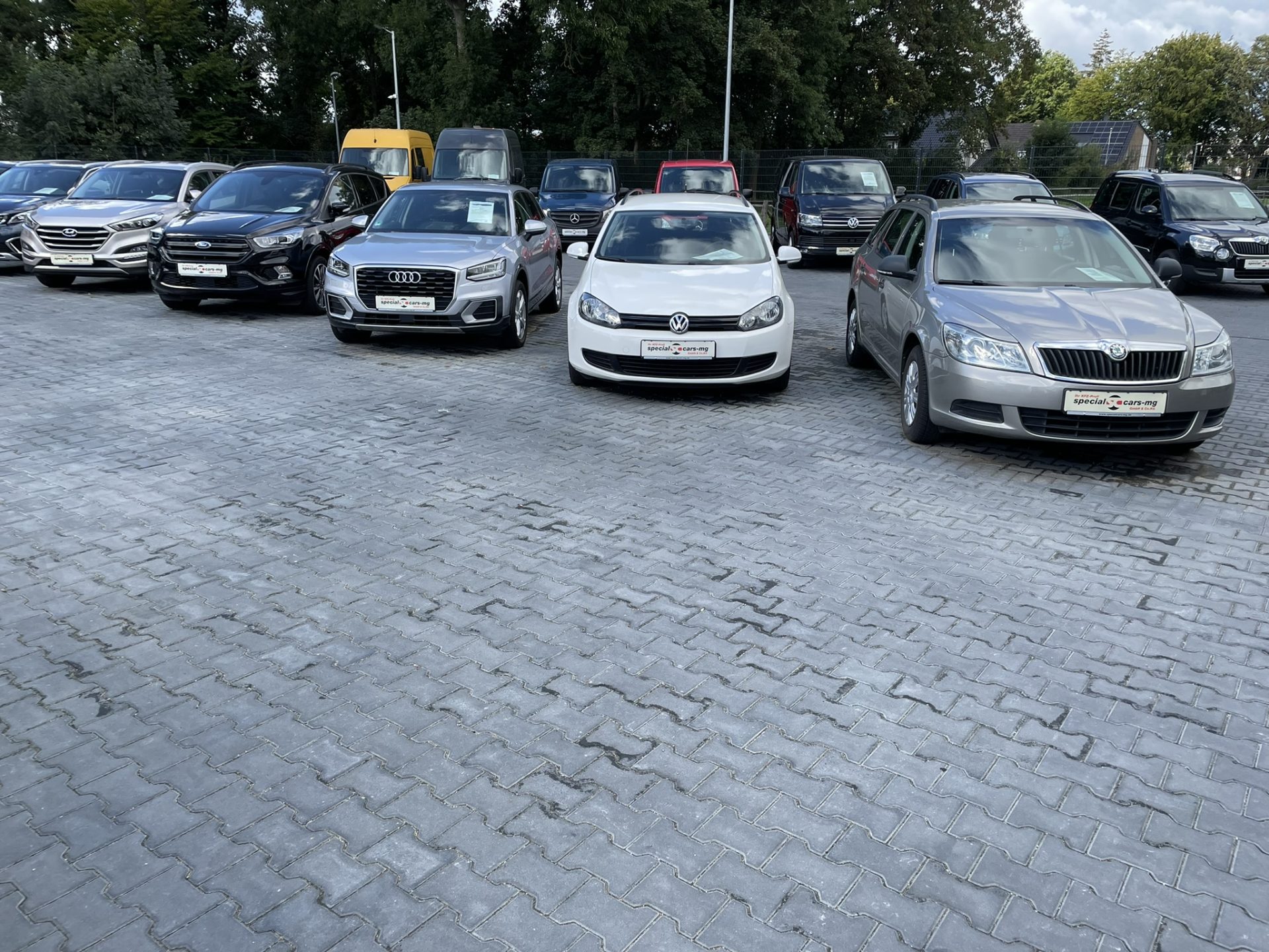 Eine Reihe verschiedener Autos, darunter SUVs und Limousinen, parkten auf einem gepflasterten Parkplatz, umgeben von Bäumen unter klarem Himmel.