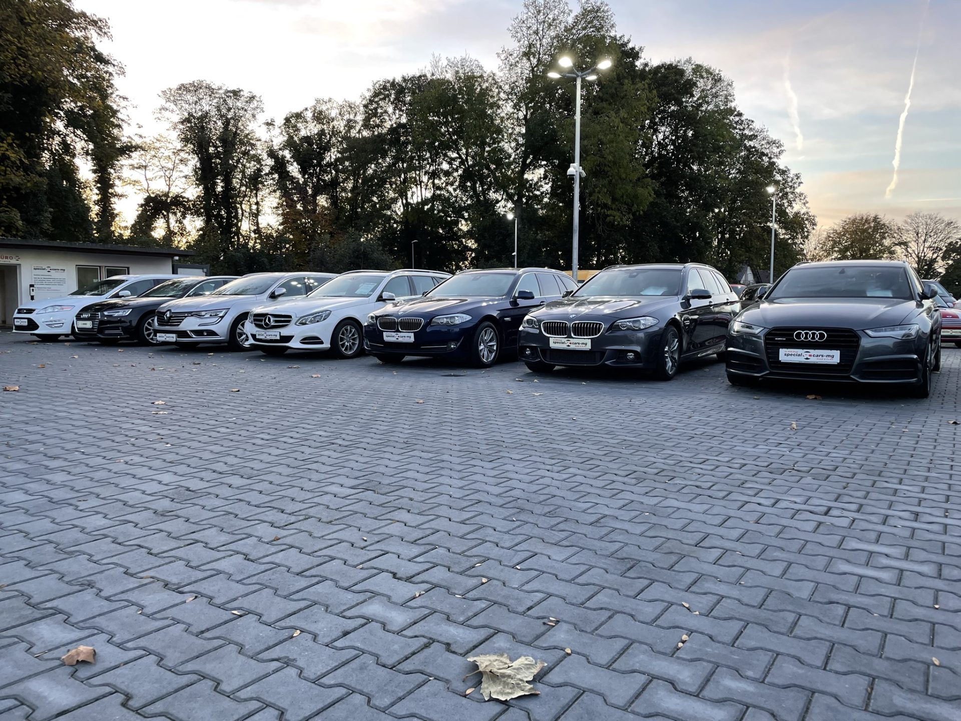 Eine Reihe von Luxusautos, darunter BMW, Audi und Mercedes, parken bei Sonnenuntergang auf einem gepflasterten Parkplatz mit Bäumen im Hintergrund.