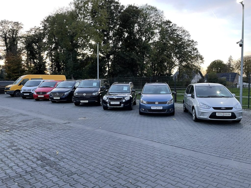 Eine Reihe verschiedener Fahrzeuge, darunter Lieferwagen und Kleinwagen, sind auf einer gepflasterten Fläche geparkt, im Hintergrund sind Bäume und ein Zaun zu sehen.