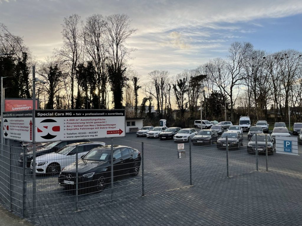 Ein Autohausgelände mit verschiedenen geparkten Autos, umgeben von Bäumen und einem auffälligen Schild mit der Aufschrift „Special Cars MG“.