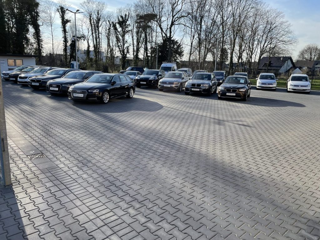 Eine Reihe verschiedener geparkter Autos, darunter Audis und BMWs, auf einem sonnenbeschienenen, von Bäumen umgebenen Parkplatz.