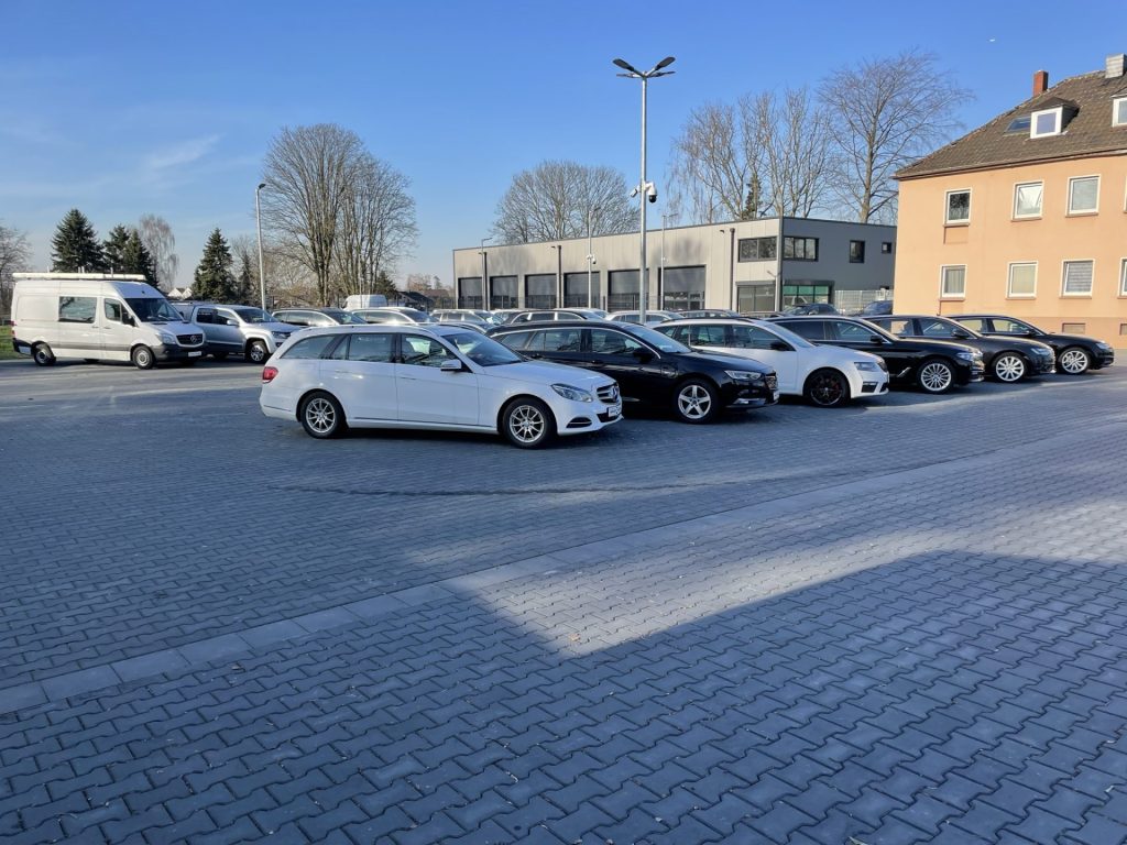 Ein sonniger Parkplatz voller verschiedener Autos, darunter ein weißer Kombi und mehrere schwarze Fahrzeuge, neben einem modernen Gebäude.