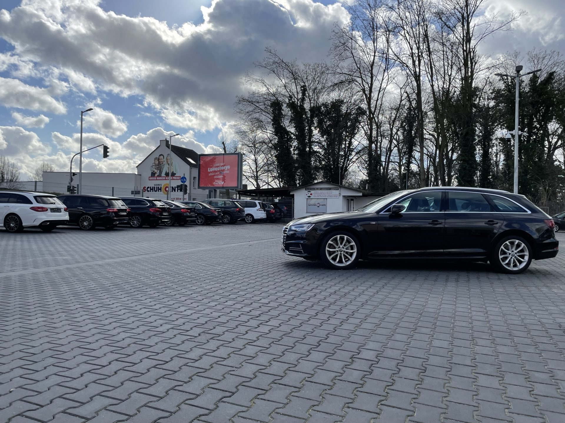 Ein schwarzes Auto parkt auf einem großen, gepflasterten Parkplatz unter einem teilweise bewölkten Himmel. Weitere Fahrzeuge stehen vor einem Gebäude mit Schildern und Bäumen aufgereiht.